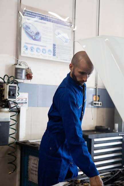 Mechanic in blue coverall checking a car engine in a modern garage setup indoors.